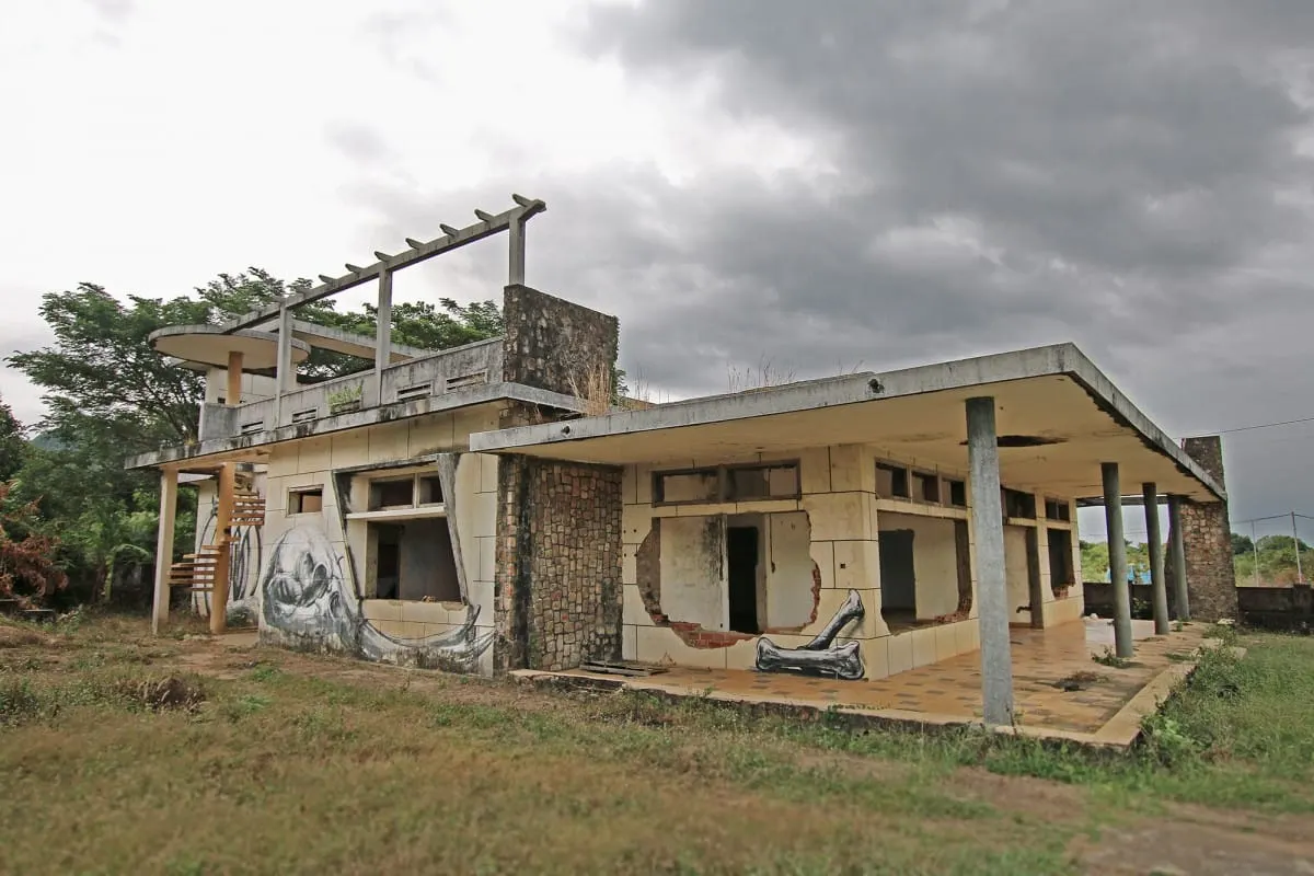 An abandoned mid-century villa with murals and broken walls under stormy skies, Kep.