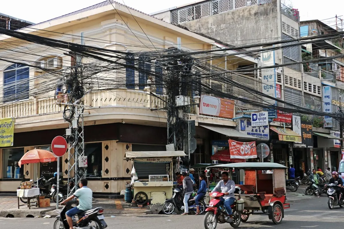 Tangled power lines over a Phnom Penh street corner with motorbikes and a tuk-tuk.