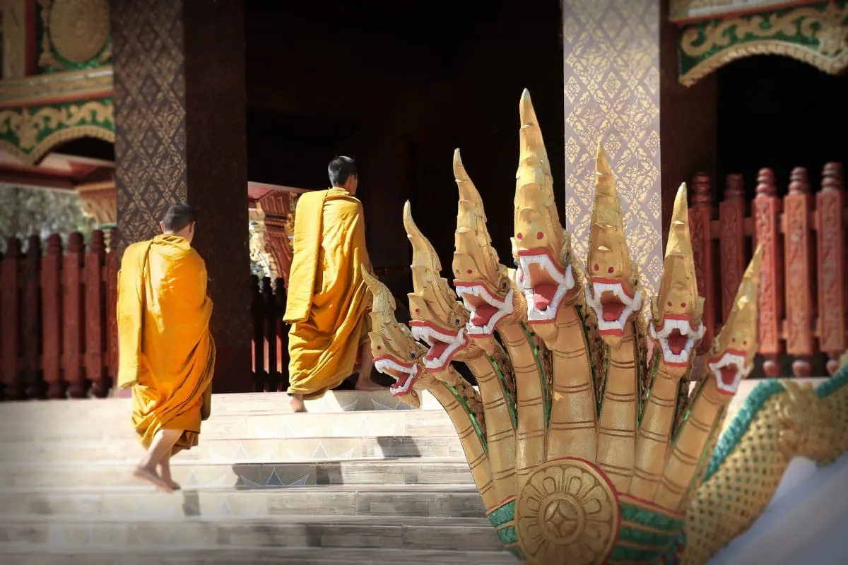Two monks in saffron robes climbing temple steps beside a golden naga statue.