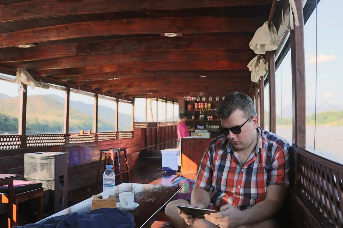 Man in sunglasses reading a tablet in the wooden cabin of a Mekong slow boat