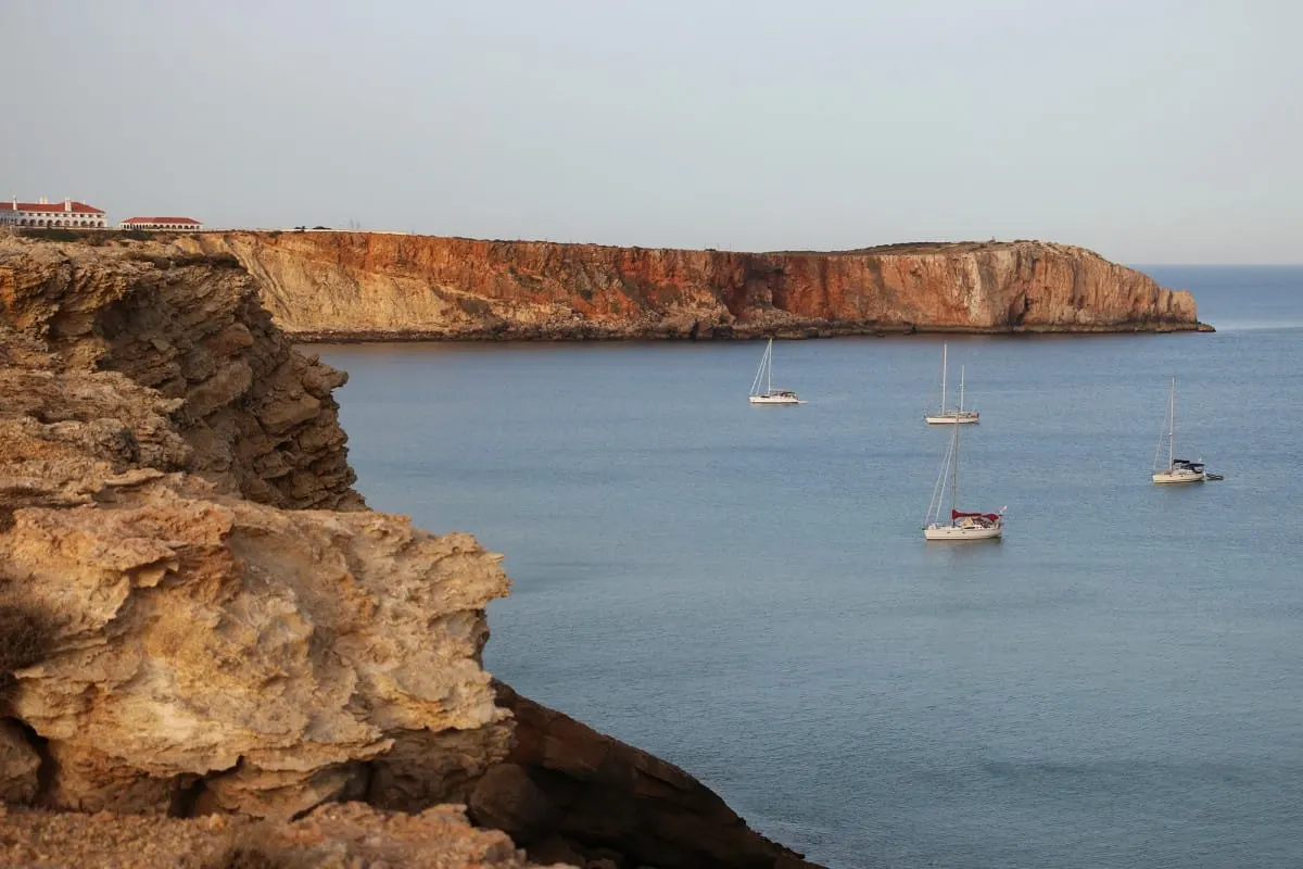 Sailboats anchored in a calm cove below ochre cliffs near Sagres