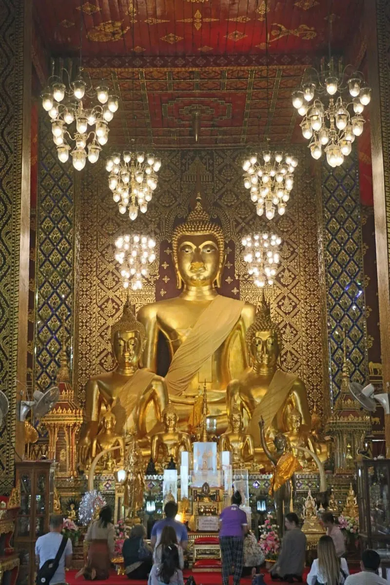 Worshippers before golden Buddha statues under chandeliers in a Lamphun temple.
