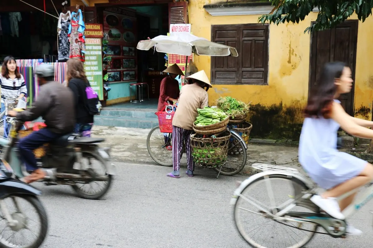 Vendors in conical hats with vegetable baskets as bikes and scooters pass